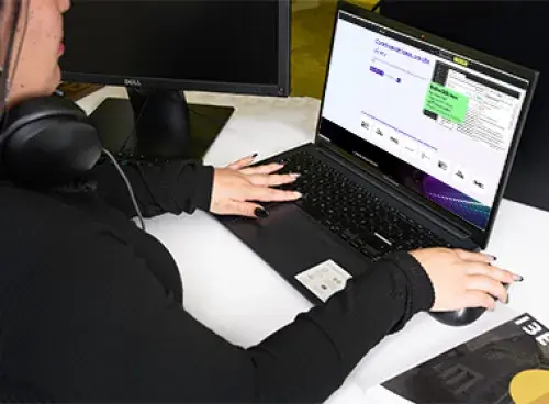 Persona frente a un portátil trabajando; a un lado hay un libro y un monitor.